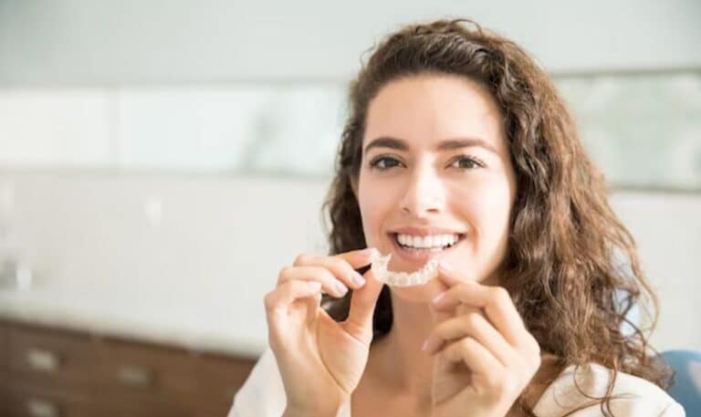 Woman smiling while holding clear aligner trays in dental office setting.