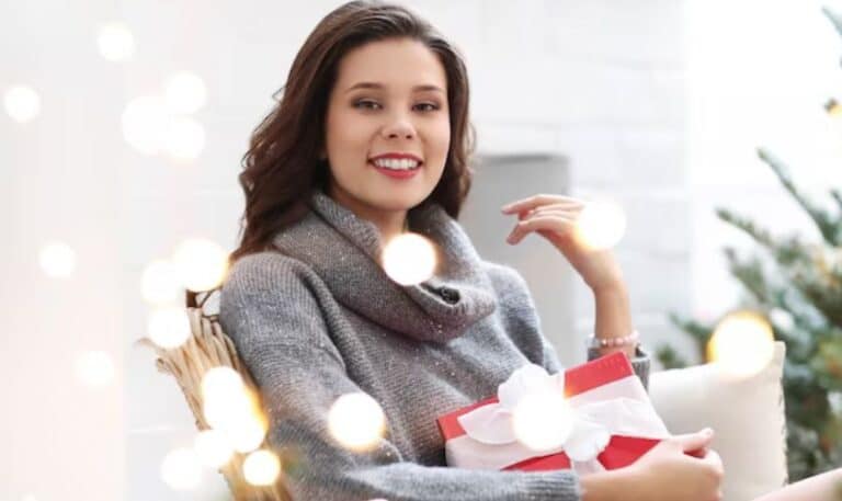 Woman holding gift box wearing gray sweater in festive holiday setting with Christmas tree.