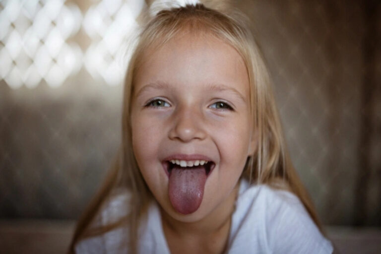 Young girl with blonde hair sticking tongue out playfully, smiling at camera indoors.