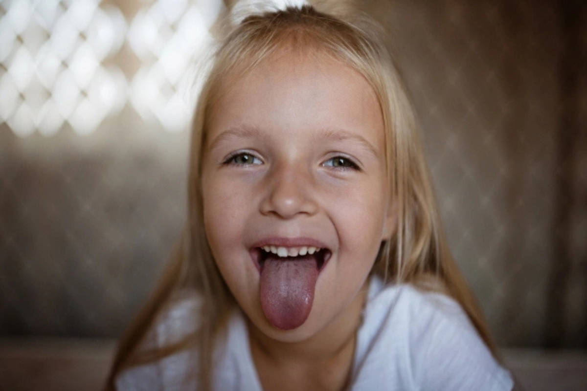 Young girl with blonde hair sticking tongue out playfully, smiling at camera indoors.